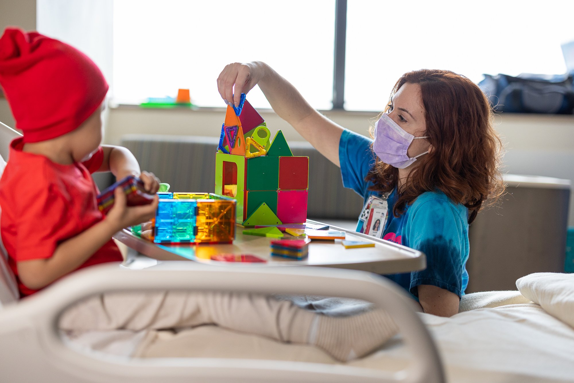 Brown-haired nurse plays with colorful magnatiles with young male patient in a red beanie