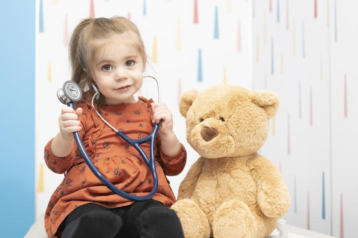 young girl holding stethoscope sitting next to stuffed bear at doctor's office