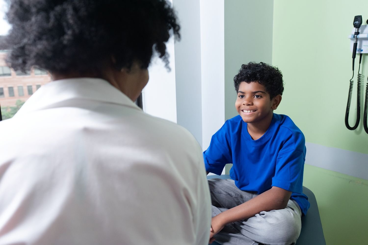 young boy in a blue shirt sitting on an exam table speaking to a provider