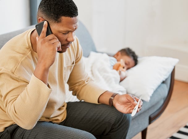 A dad sits on a couch on the phone while he looks at a thermometer with his son's temperature as his son lays beside him.