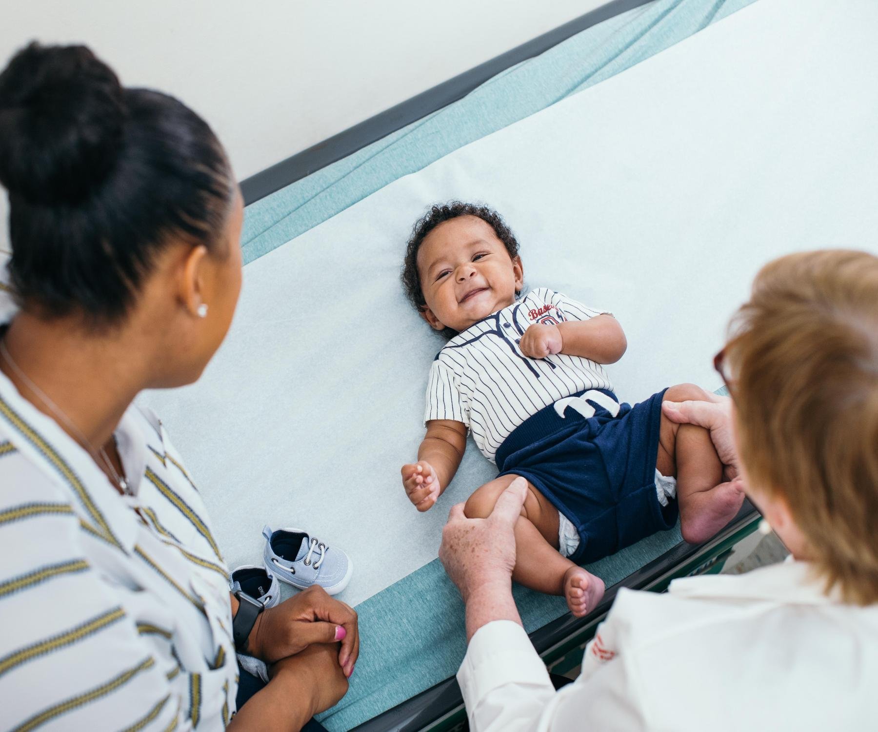 smiling baby on exam table with mother and provider