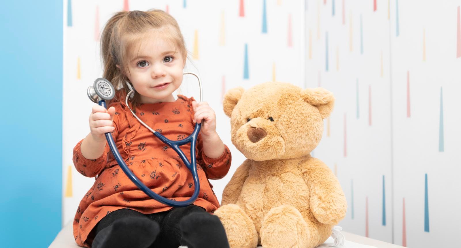 blonde girl holding stethoscope on exam table next to teddy bear