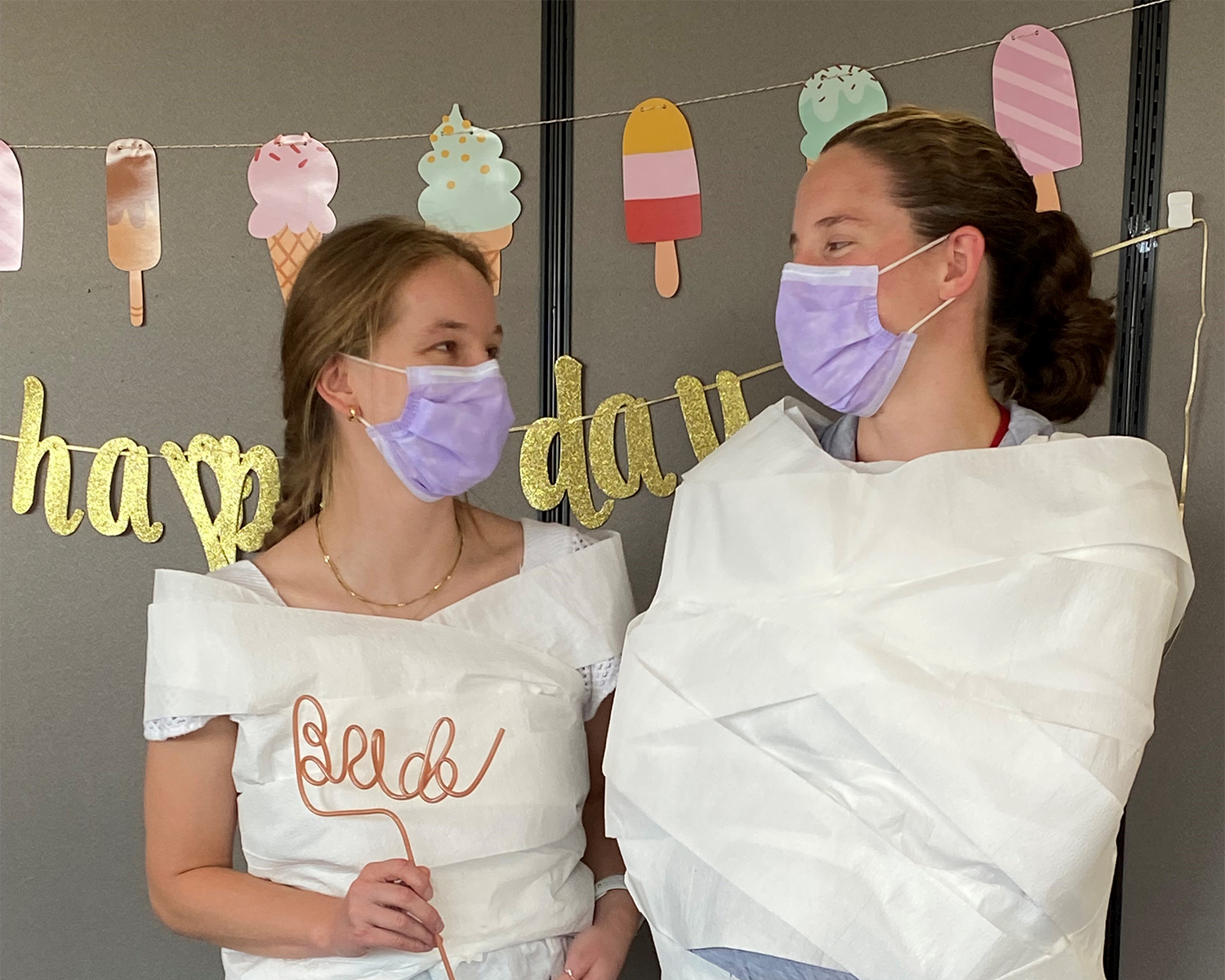 Two women wearing masks and one of them wrapped in toilet paper from a game