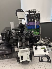 Black and white confocal inverted microscope stand. Laser unit behind with purple, blue dark green, light green stacked boxes.