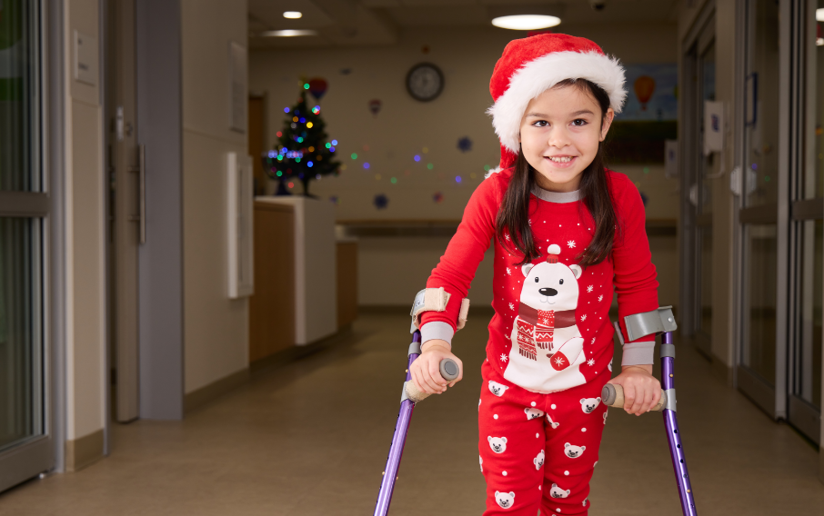 A young female patient on crutches smiles at the camera while wearing red winter pajamas and a Santa Claus hat