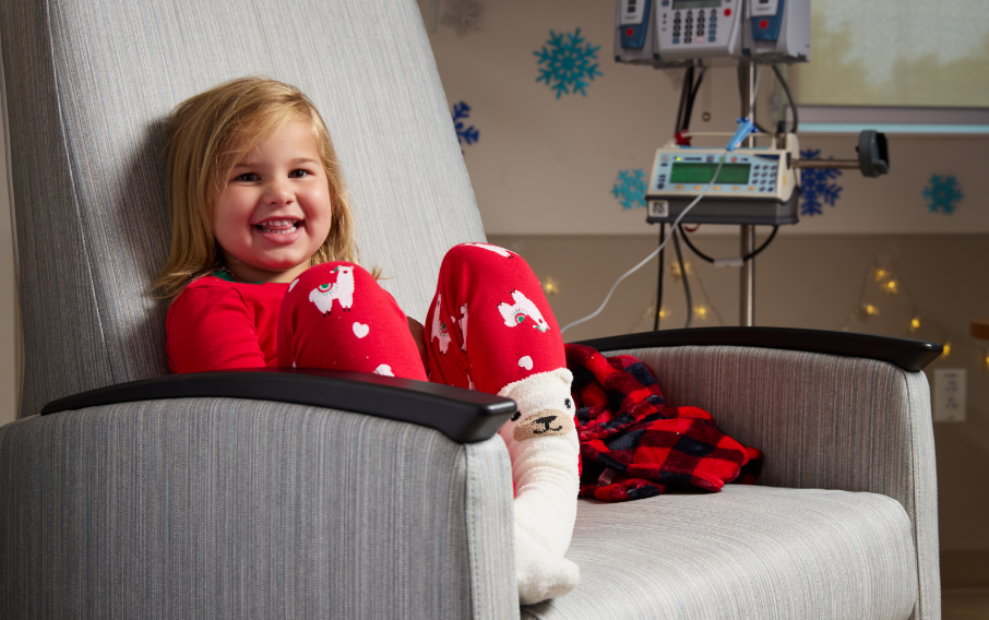 A young patient smiles at the camera while sitting in a chair next to a monitoring device