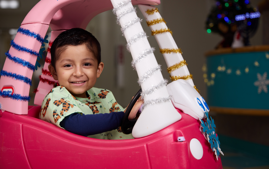 A young patient smiles at the camera while pretending to drive a miniature car decorated with tinsel and snowflakes