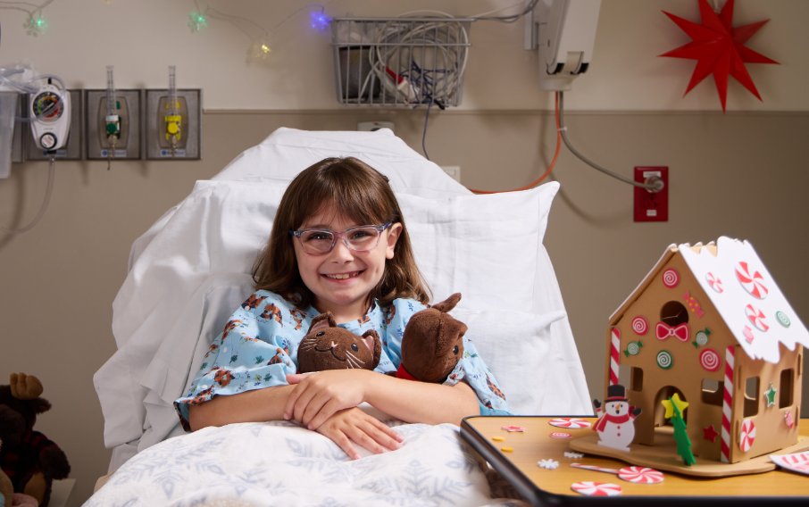 A young patient smiles at the camera while reclining in a bed next to a gingerbread house art project