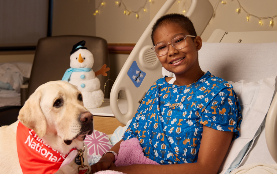 A patient smiles at the camera while reclined in a bed and posing with a therapy dog