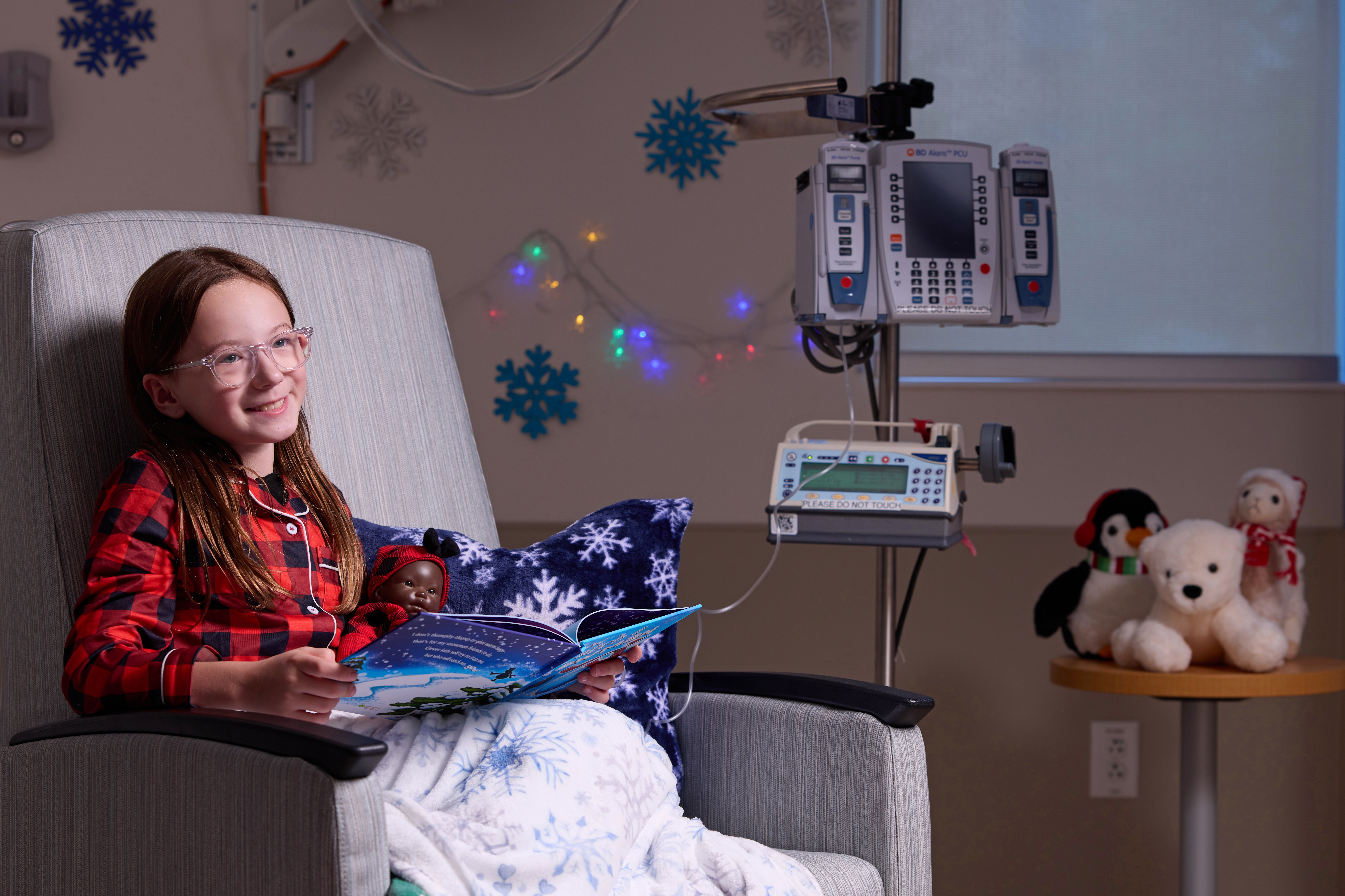 Patient McKenzie sits in a lounge chair and reads to a baby doll while wearing matching flannel outfits.