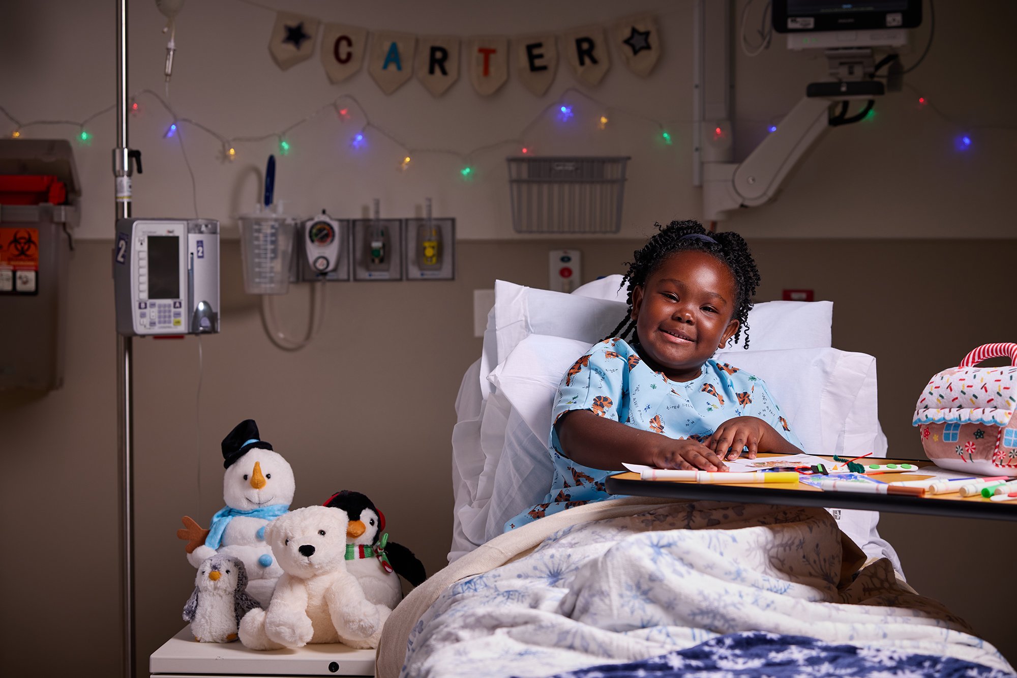 CNH patient Carter, age 6, smiles at the camera while sitting in a hospital bed surrounded by stuffed animals.