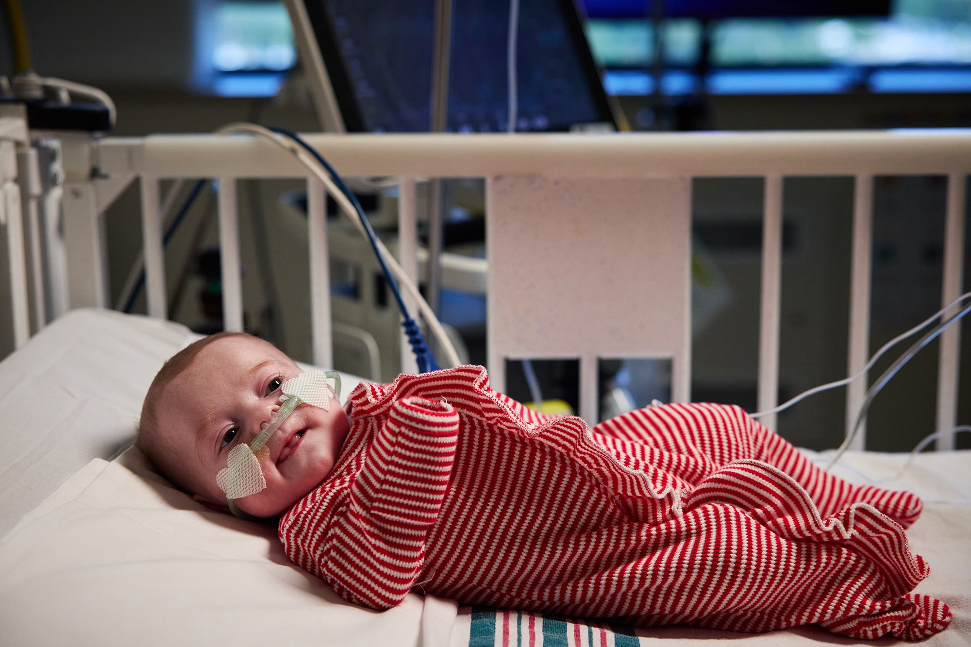 CNH patient Eloise, age 3 months, laying in a bed and receiving supplemental oxygen while wearing a red and white flannel onesie.
