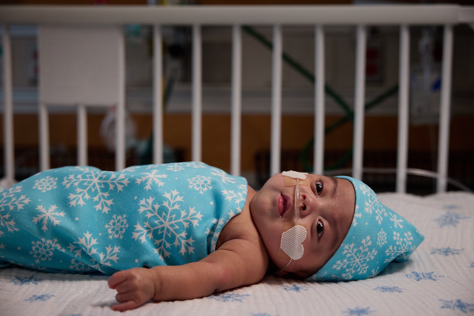 CNH patient Mateo, age 8 months, lays in a crib swaddled in a snowflake blanket with matching hat.