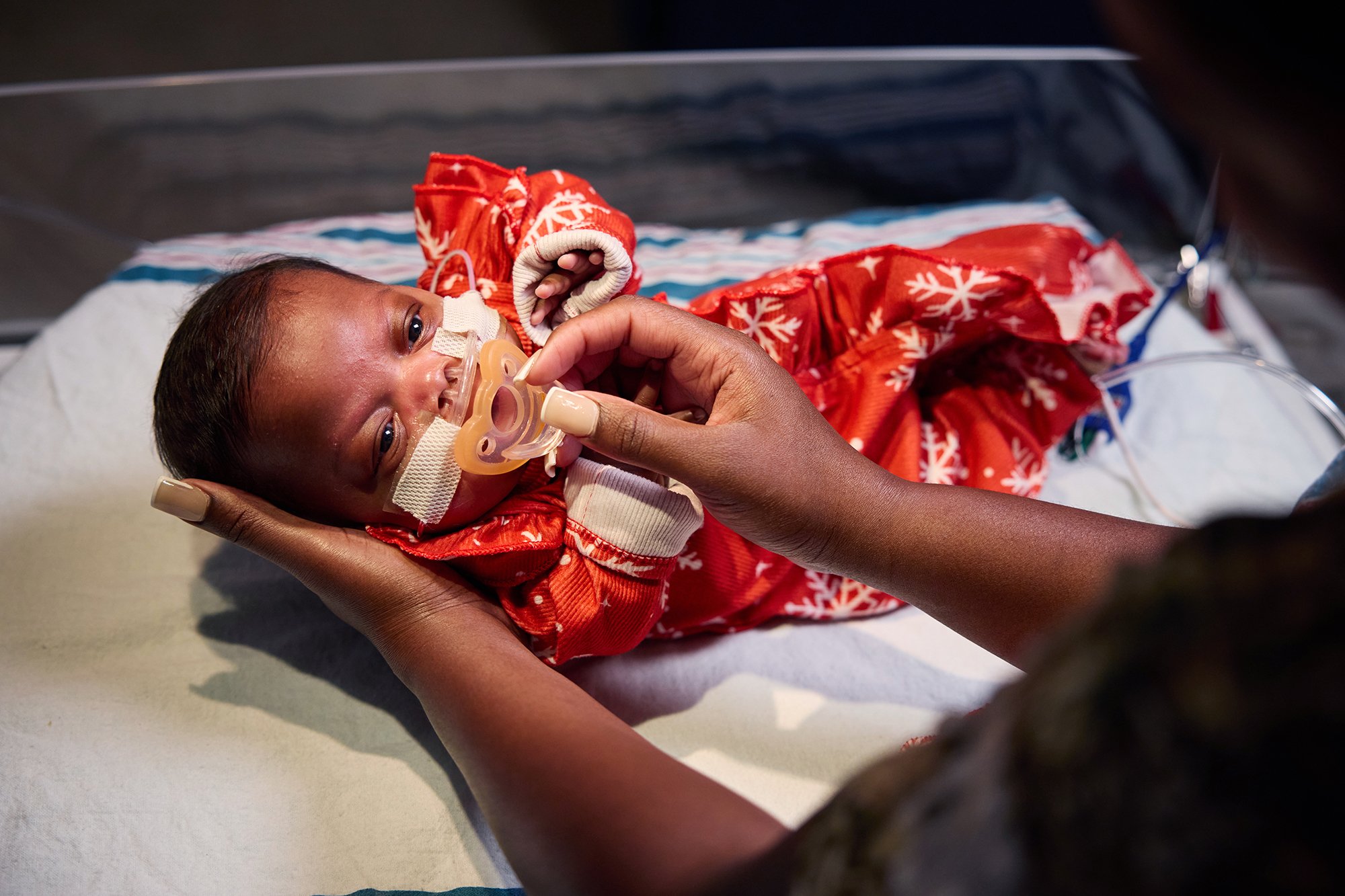 CNH patient Na'Kahri, age 3 months, looks at a caregiver while laying in a hospital bassinet and wearing a snowflake onesie.