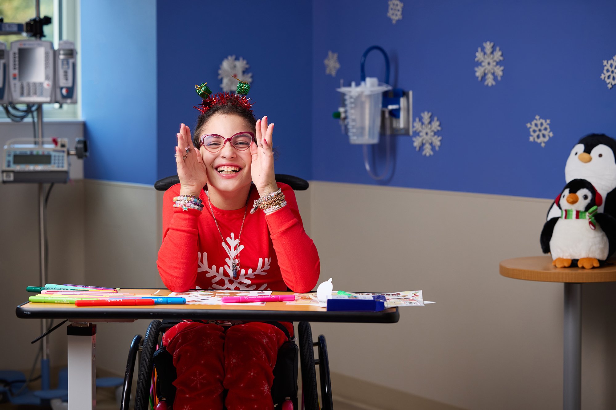CNH patient Salma, age 10, sits in a wheelchair and smiles at the camera while making art in snowflake-themed pajamas.