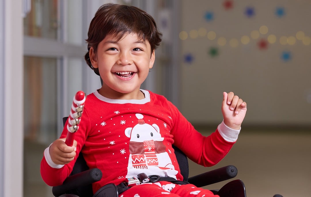 Uly, a young male patient, sits in a wheelchair and smiles while wearing holiday-themed pajamas.