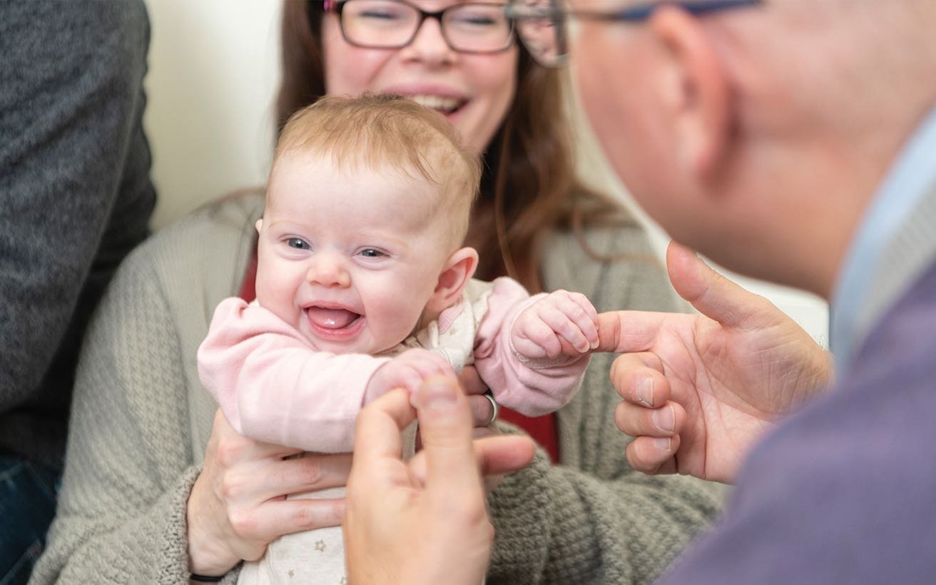 A White, female infant smiles in her mother's lap while holding the hands of a White, male surgeon.