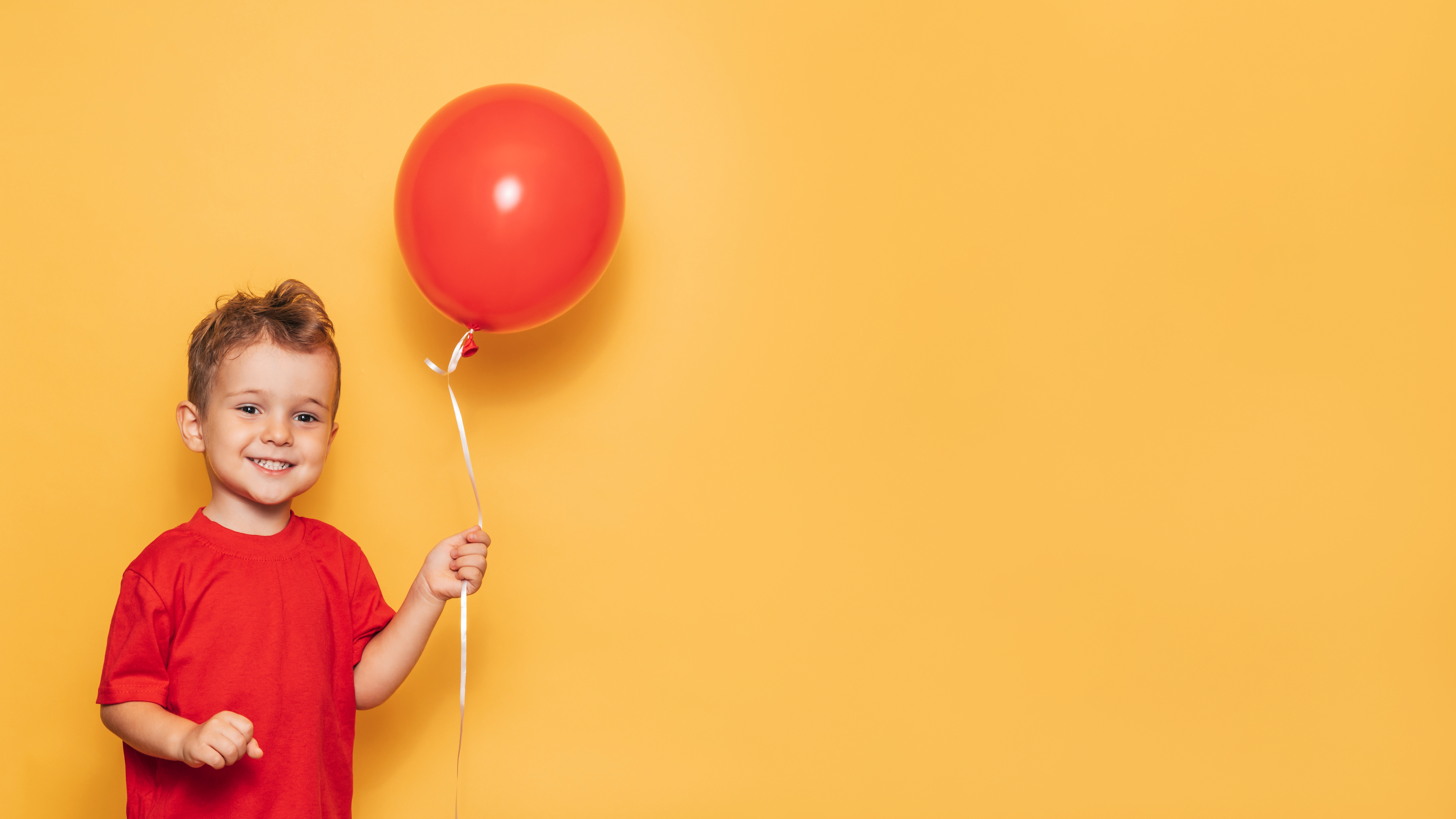 A happy Caucasian boy isolated on a bright yellow background holds a red balloon in his hands.