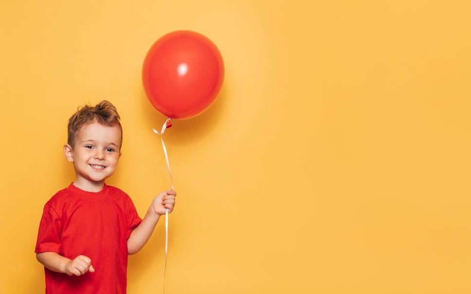 A happy Caucasian boy isolated on a bright yellow background holds a red balloon in his hands.