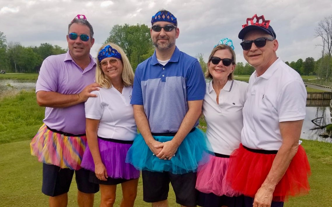 Five adults in golf gear and tutus pose for a photo on a golf course.