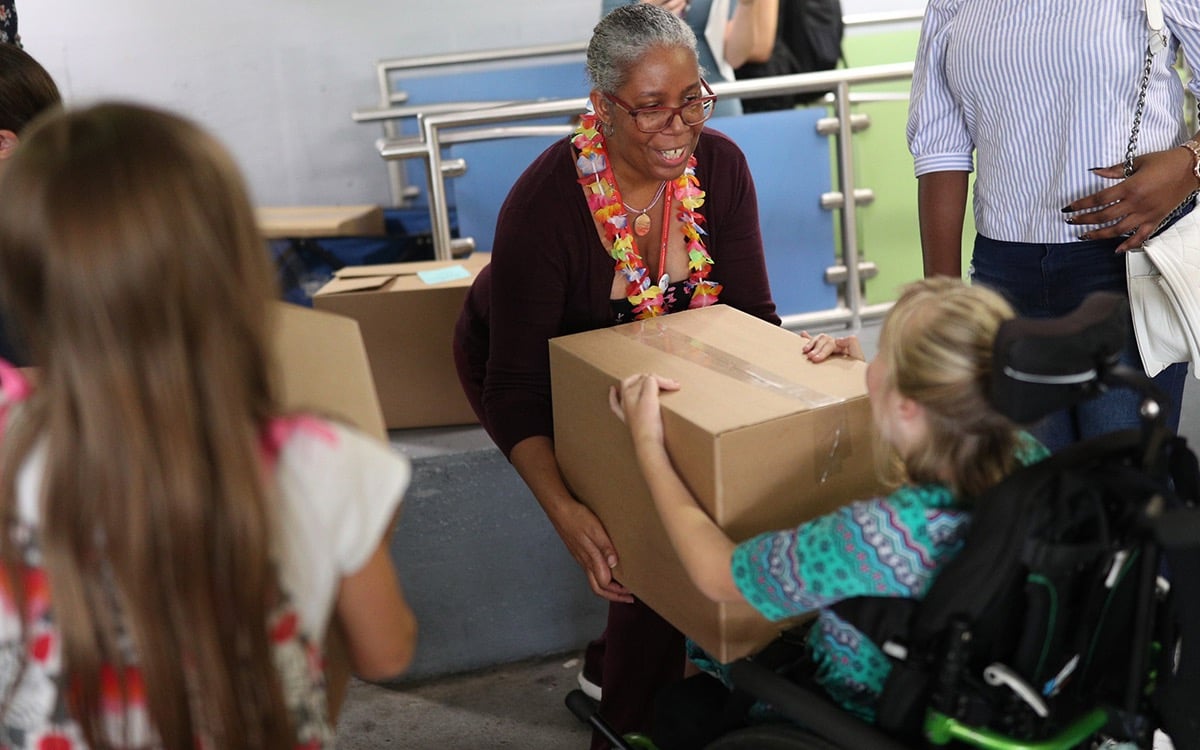 A smiling woman hands a box to a young person in a wheelchair.