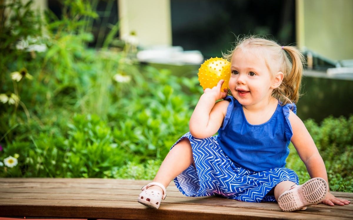 Female toddler sitting on a bench and holding a yellow ball.