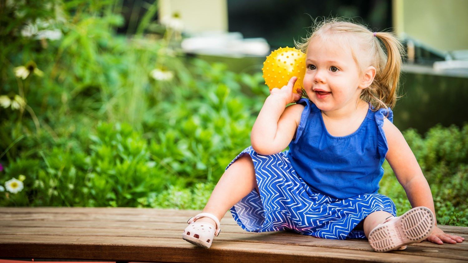 Female toddler sitting on a bench and holding a yellow ball.