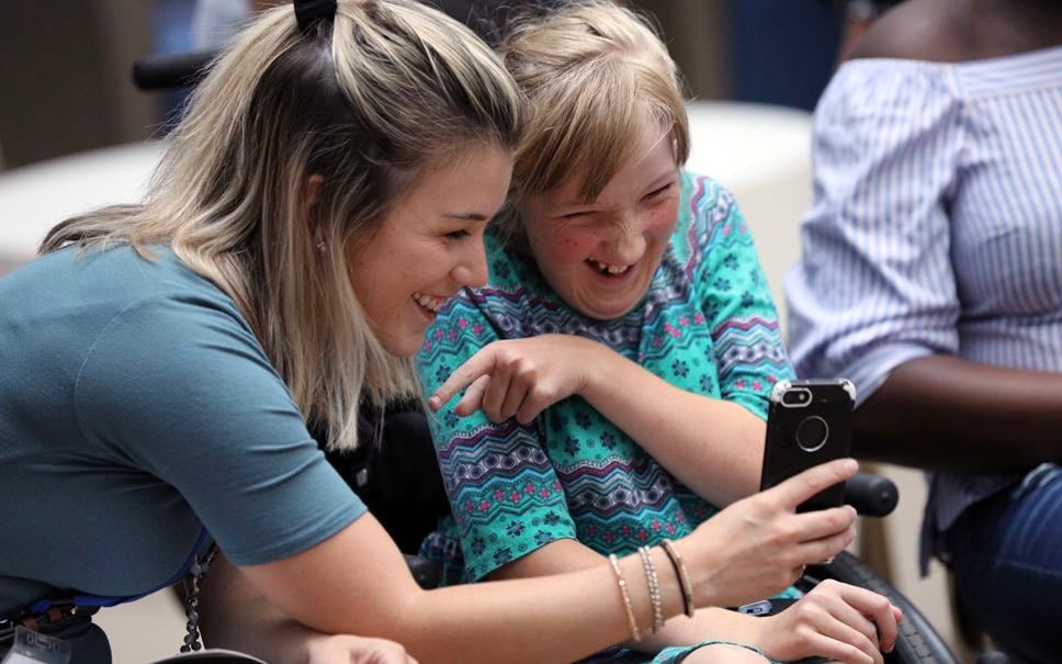 A White female volunteer and a White female patient in a wheelchair laugh as they take a selfie.