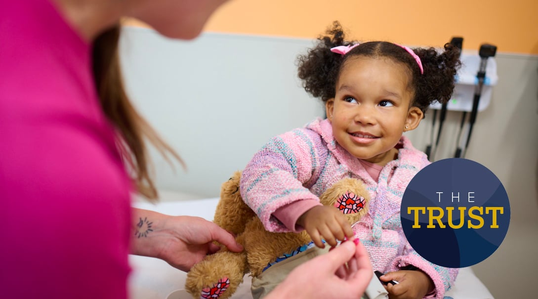 The Trust logo overlaid on an image of a medical provider offering a stuffed animal toy to a young female patient