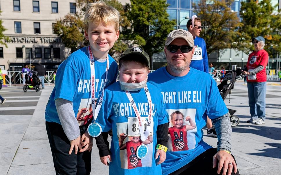 A father poses with his two young sons, one of whom is a Children's National patient. This White family are wearing matching t-shirts that say, "Fight like Eli!"