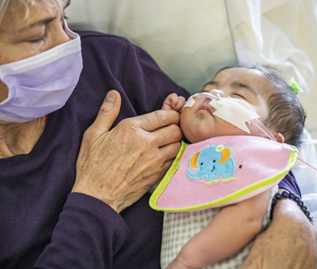 A woman wearing a medical mask cradles a young infant breathing supplemental oxygen.