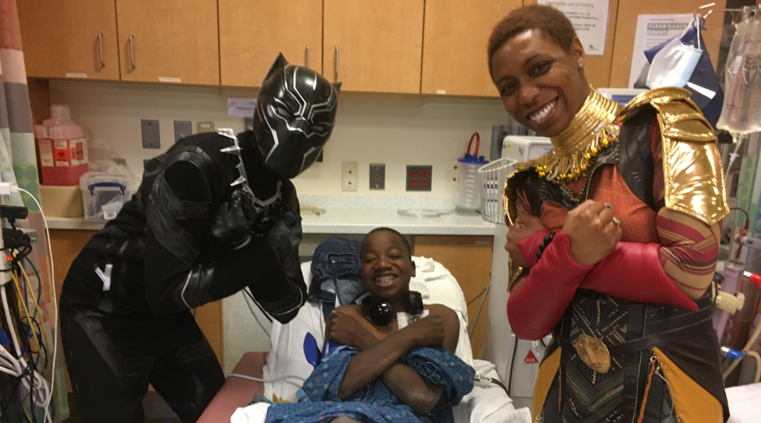 Caleb, a young male patient, in a reclined position making the Wakanda Forever salute alongside characters from the Black Panther movie
