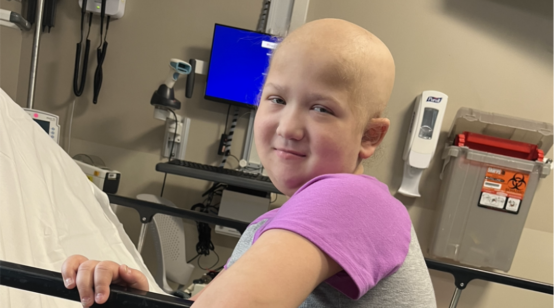 Charlotte, a young female bald patient, sitting on a hospital bed and smiling at the camera during treatment