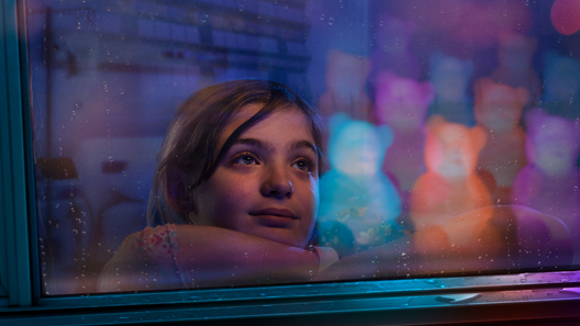 An elementary school aged female patient looks longingly out a window with the glow of Light Up Dr. Bears in the reflection.