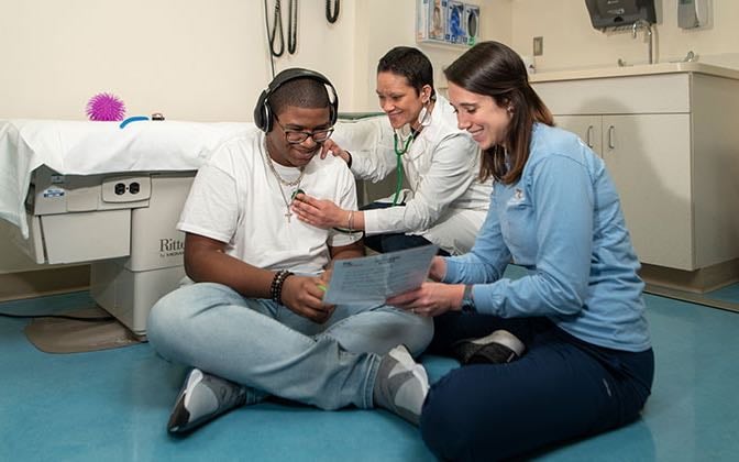 A teen with autism sits cross-legged on the floor as their heartbeat is checked and they work with a therapist.