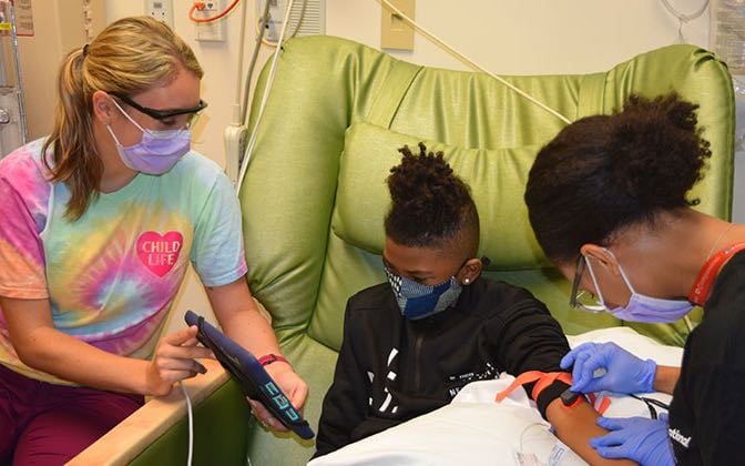 Two female nurses, one Black and one White, attend to a Black elementary school aged boy. One nurse holds an iPad so the patient can video chat with family.