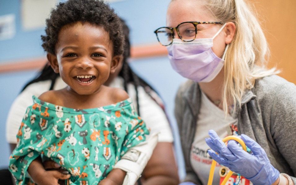 A pre-school aged Black male patient smiles in his colorful hospital gown along side a White female healthcare professional.