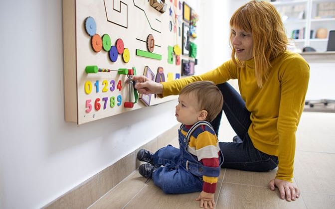 A mother and her baby sit on the floor and play with a wall-mounted activity puzzle.