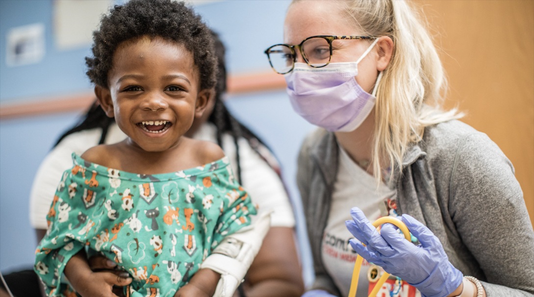 A pre-school aged male Black patient smiles alongside a Children's National healthcare professional.