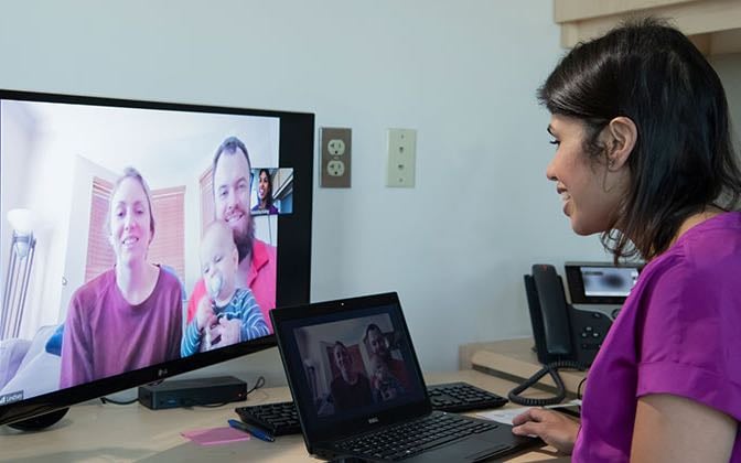 A White family is shown on a monitor, and a female practitioner of color speaks with them via video chat from their office.