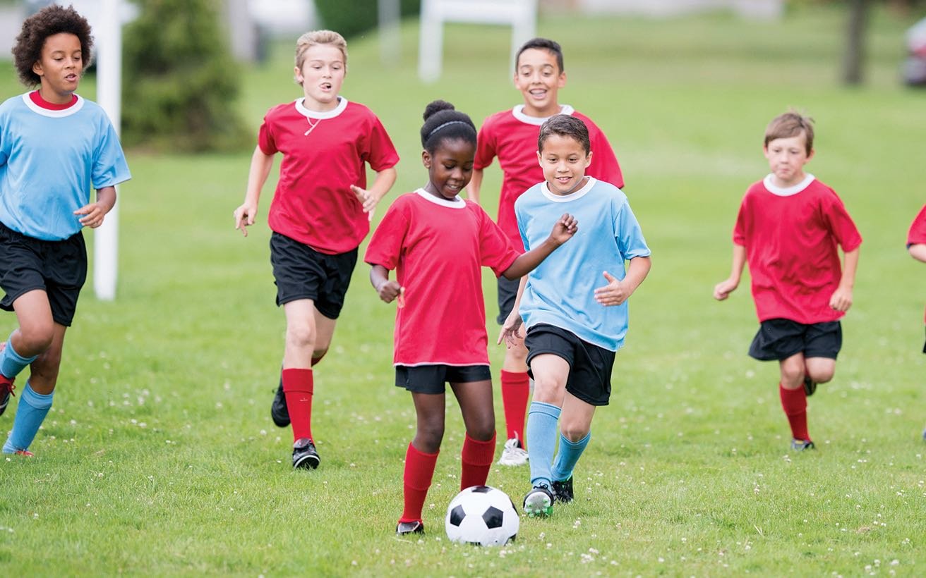 A group of children of multiple genders, ages, and races play soccer together.