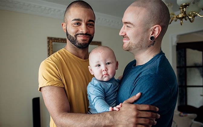 Two dads embrace holding their infant child.