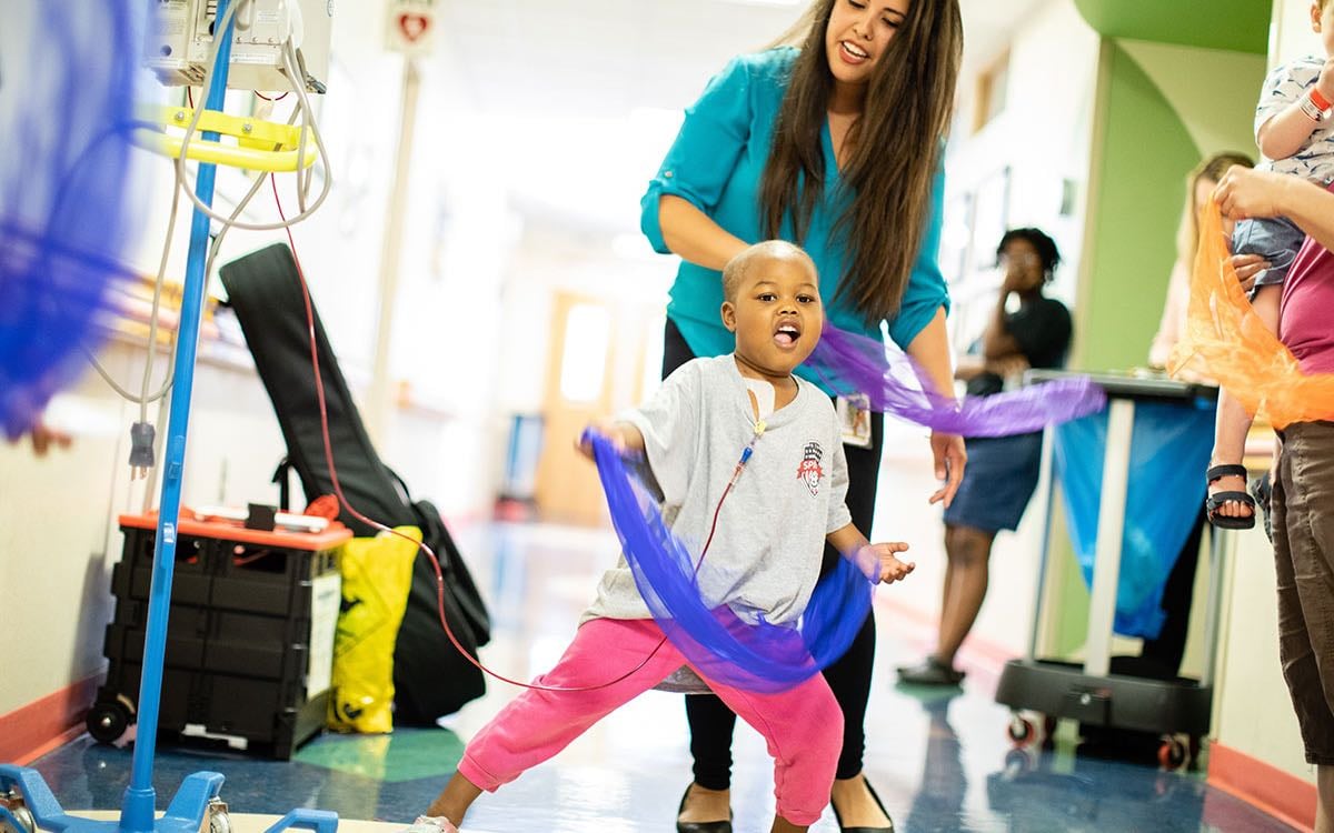 An early school-aged patient waves a scarf as they dance in a hospital hallway.