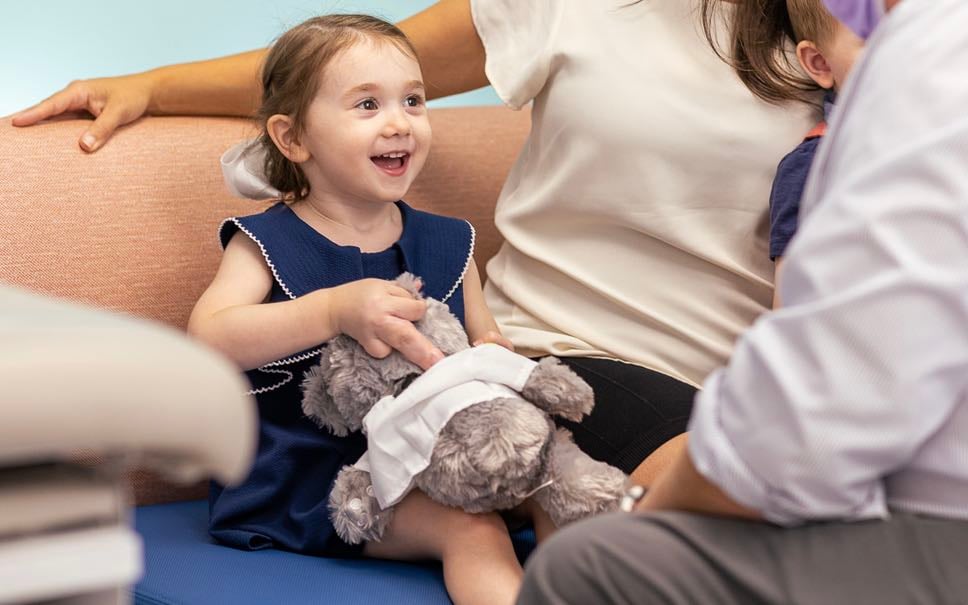 A pre-school aged White female patient holds a Dr. Bear doll while a doctor speaks with her and her mother.