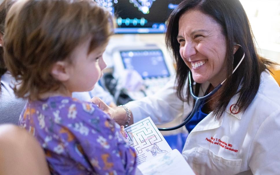 Dr. Donofrio, a White female physician, smiles as she confers with a toddler-aged White female patient.