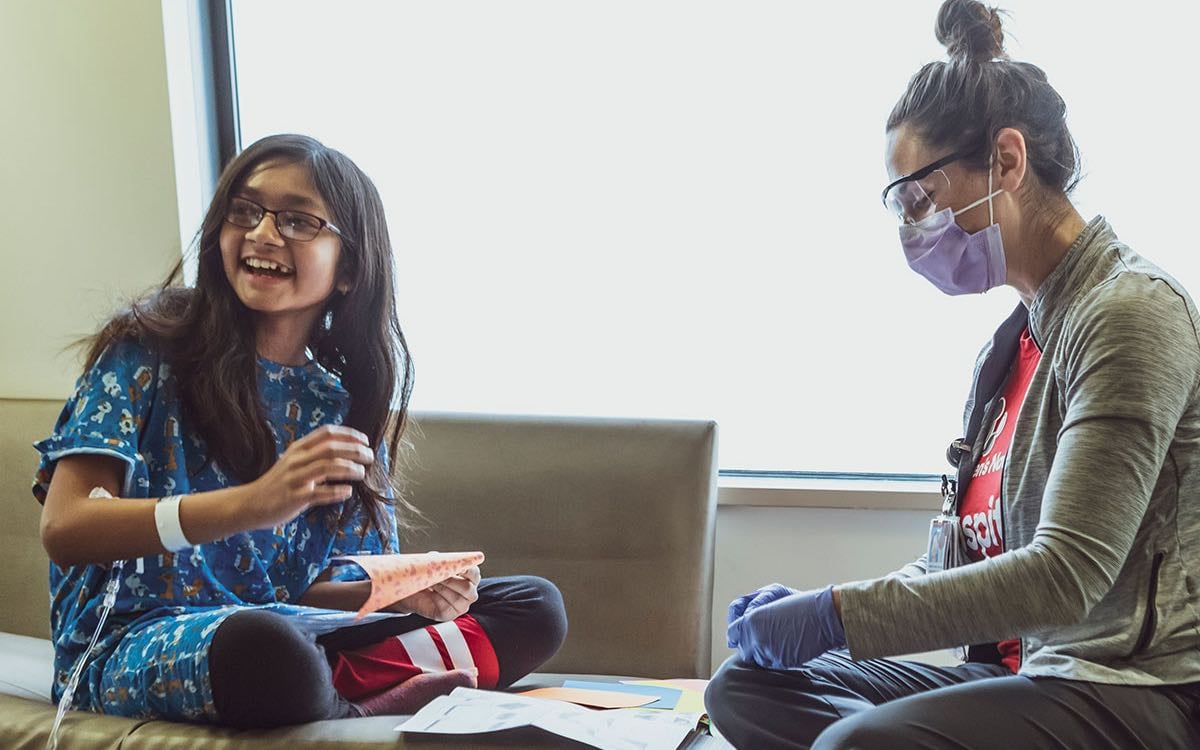 A young patient laughs as she works on homework and sits with her nurse.