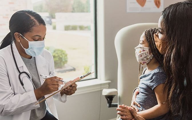 A doctor evaluates a young patient who is sitting on their mother's lap in an exam room.