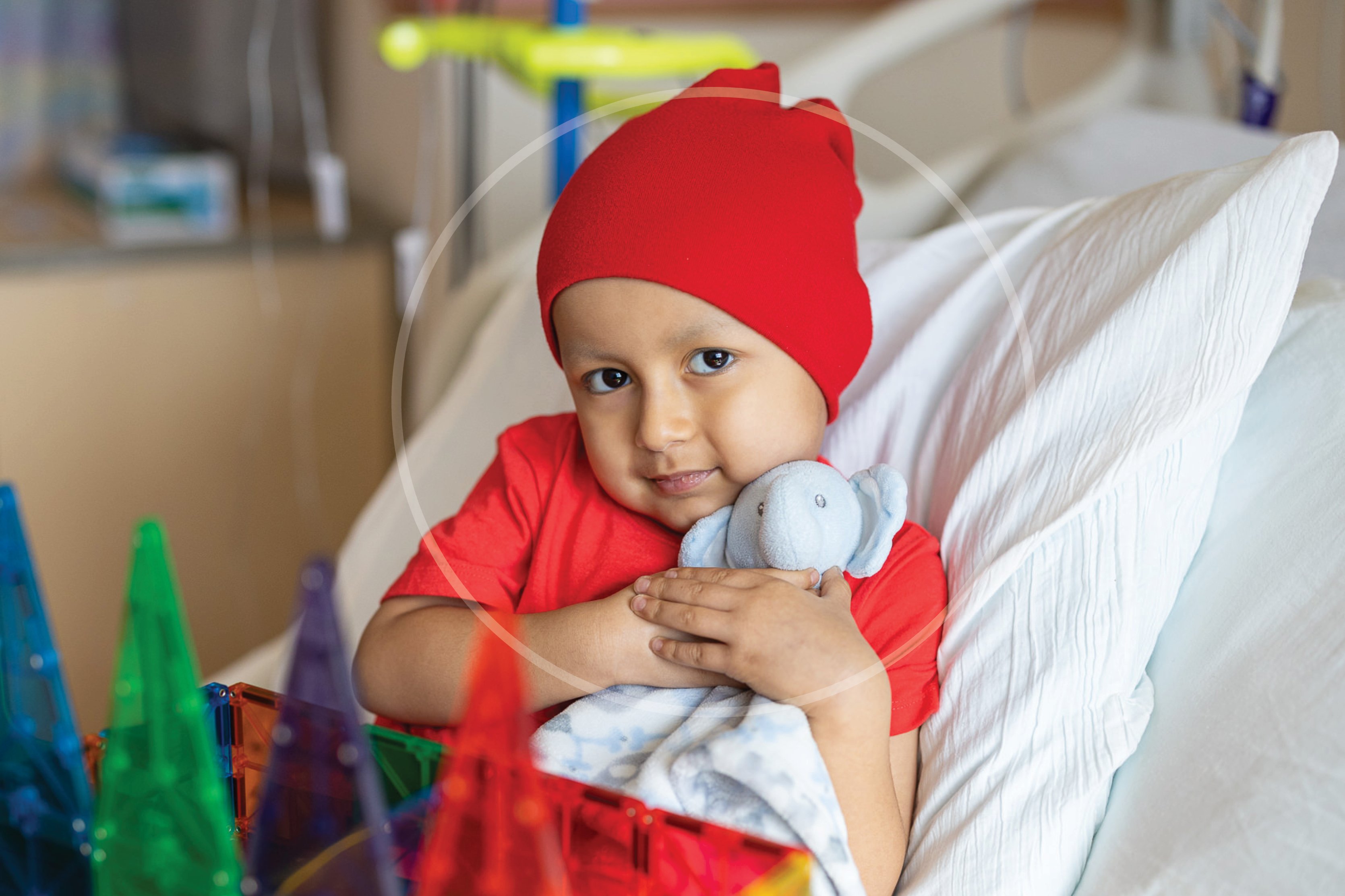 A young child of color in a hospital bed wearing a red cap hugging a elephant stuffed animal