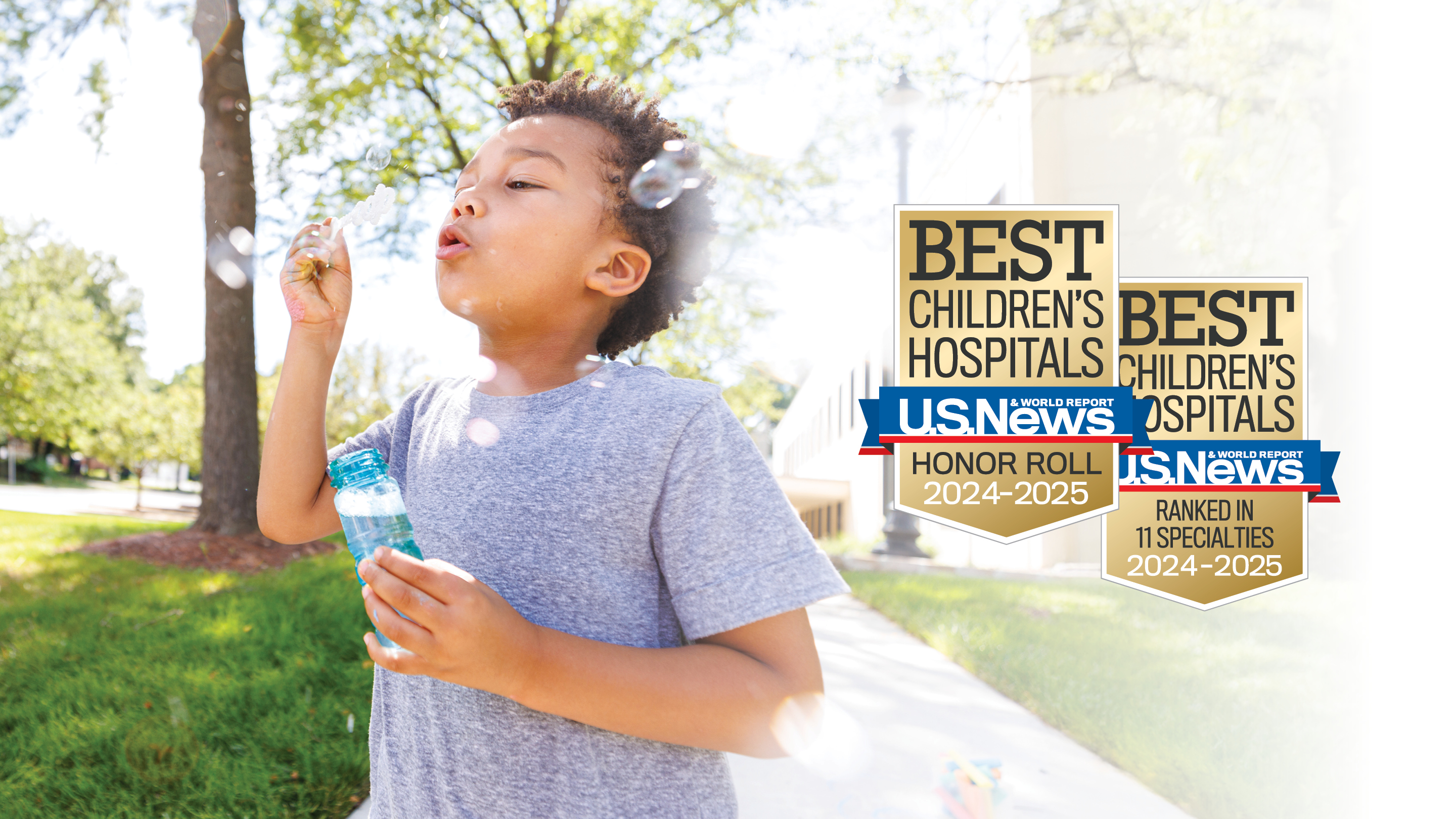 A boy of color is outside by trees blowing bubbles with US News Honor Roll badges