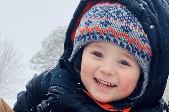 A toddler dressed in a snowsuit smiles in his parent's arms.